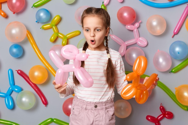 Verbaasd kind Verrast feestelijk meisje met vlechten die tegen grijze muur staan en kleurrijke ballonnen vasthouden die met grote ogen en open mond verbazing uiten
