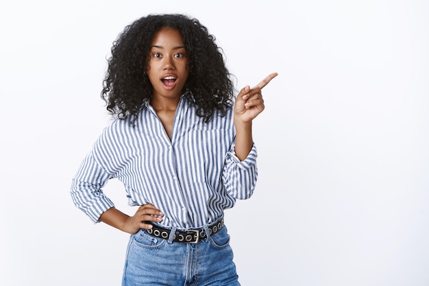 Verbaasd onder de indruk vrouw vraagt vriend wat was dat zijwaarts wijzend nieuwsgierig wijzend wijzend wijsvinger open mond geïntrigeerd terloops terloops hand taille, gestreepte blouse dragen