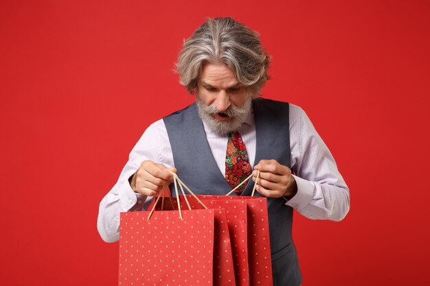 Verbaasd oudere grijsharige snor bebaarde man in overhemd vest stropdas poseren geïsoleerd op rode achtergrond. Mensen levensstijl concept. Bespotten kopie ruimte. Pakkettas vasthouden met aankopen na het winkelen.