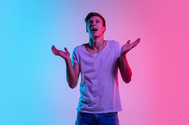 Verbaasd. Portret van een jonge blanke man op de achtergrond van de gradiënt blauw-roze studio in neonlicht. Concept van jeugd, menselijke emoties, gezichtsuitdrukking, verkoop, advertentie. Mooi model in casual.