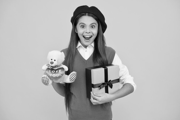 Verbaasd tienermeisje in Franse baret houdt speelgoed vast en geschenkdoos op gele achtergrond verbazing