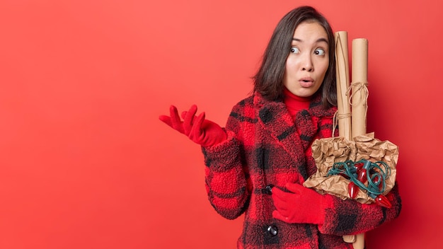 Verbaasd verbaasd Aziatische vrouw haalt schouders op, voelt aarzelend kijkt weg onder de indruk poses met nieuwjaarsattributen gekleed in bovenkleding geïsoleerd op rode achtergrond blanco kopie ruimte voor uw tekst