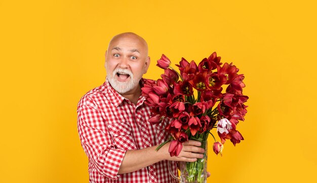 Verbaasd volwassen gepensioneerde man met baard houden tulpen lentebloemen op gele achtergrond