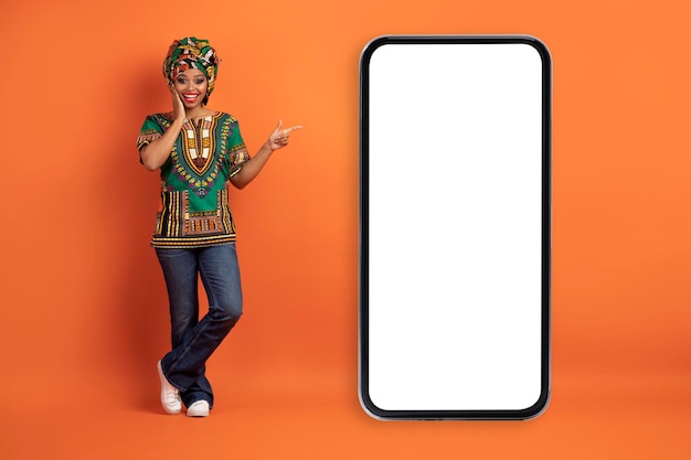 Verbaasde Afrikaanse vrouw wijzend op enorme mobiele telefoon oranje achtergrond