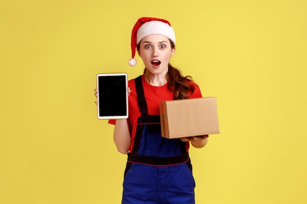Verbaasde bezorger die tablet met leeg scherm toont voor promotie en pakket vasthoudt met blauwe overalls en kerstman hoed indoor studio shot geïsoleerd op gele achtergrond