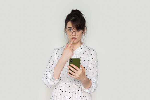 Verbaasde blanke brunette vrouw raakt haar lippen aan terwijl ze een telefoon op een witte muur houdt