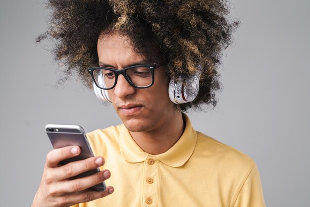 verbaasde blanke man met afro-kapsel met koptelefoon en bril met smartphone geïsoleerd over grijze muur