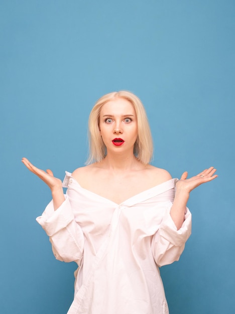 Verbaasde blonde vrouw in wit oversized shirt geïsoleerd op blauwe achtergrond op zoek naar camera