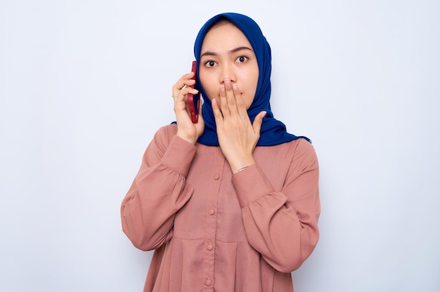 Verbaasde jonge Aziatische vrouw in roze shirt praten op mobiele telefoon en die betrekking hebben op mond geïsoleerd op witte achtergrond Mensen religieuze levensstijl concept