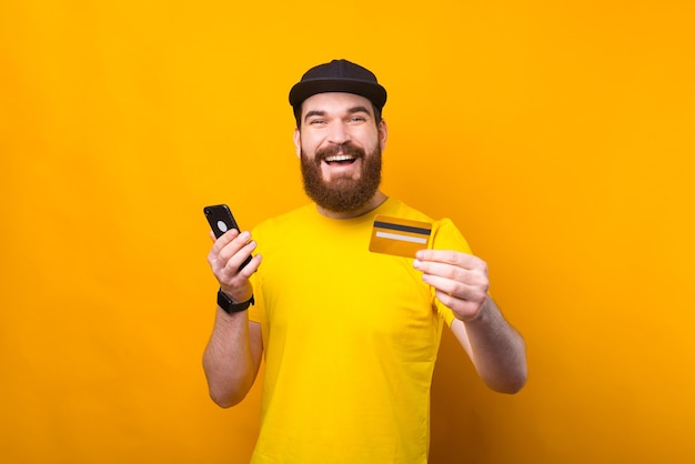 Verbaasde jonge bebaarde man met creditcard en smartphone op gele achtergrond