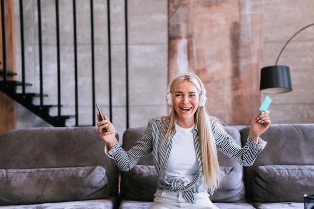 Verbaasde jonge blonde Zweedse vrouw met telefoon en creditcard in koptelefoon zittend op gezellige bank schreeuwend gelukkig opgeheven handen Online aankoop korting verkoop Black Friday Succesvolle mensen