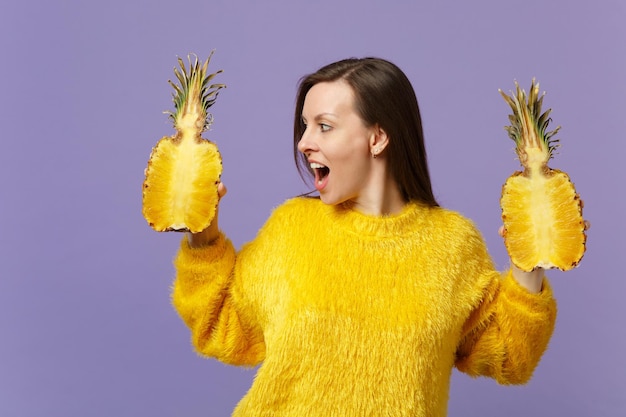 Verbaasde jonge vrouw die opzij kijkt en de mond openhoudt, houdt de helft van vers rijp ananasfruit geïsoleerd op een violette pastelachtergrond. Mensen levendige levensstijl, ontspannen vakantie concept. Bespotten kopie ruimte.