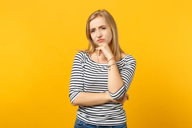 Verbaasde jonge vrouw in gestreepte kleding op zoek naar camera, zet handsteun op kin geïsoleerd op geeloranje muurachtergrond in studio. Mensen oprechte emoties, lifestyle concept. Bespotten kopie ruimte.