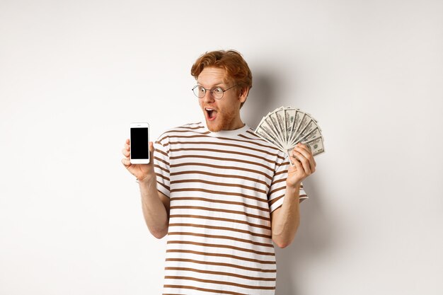 Verbaasde roodharige man smartphone app tonen op leeg scherm en geld, prijzengeld online winnen, staande op witte achtergrond.