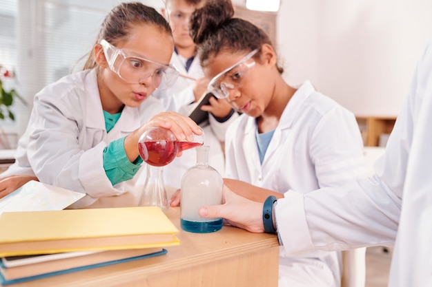 Verbaasde schoolmeisjes in beschermende brillen en witte jassen kijken naar blauwe vloeibare substantie in grote snavel vastgehouden door hun scheikundeleraar tijdens les