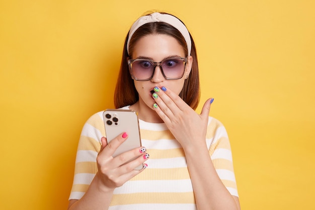 Verbaasde verbaasde jonge blanke vrouw met gestreept t-shirt en zonnebril poseren geïsoleerd over gele achtergrond permanent met mobiele telefoon schokkend nieuws lezen die mond met palm bedekt