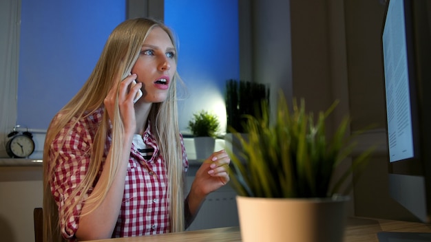 Verbaasde vrouw die op computer op smartphone spreekt. Aantrekkelijke blonde vrouwelijke vergadering op houten bureau en kijken met open mond op computermonitor praten op mobiele telefoon in de donkere kamer 's nachts