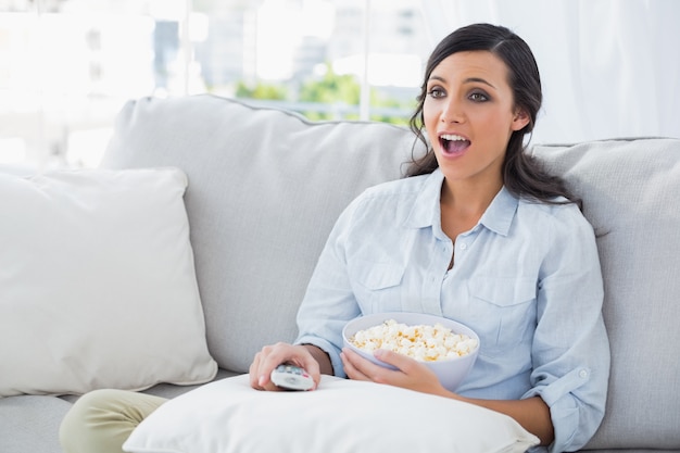 Verbaasde vrouw die op de bank ontspannen die popcorn eet