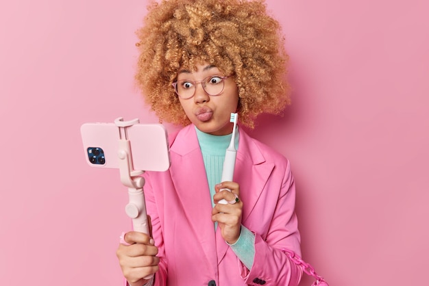 Foto verbaasde vrouwelijke blogger legt uit hoe je een innovatieve elektrische tandenborstel gebruikt, houdt de lippen afgerond, draagt een transparante bril en een formele jas poseert bij de camera aan de voorkant van de mobiele telefoon op selfiestick