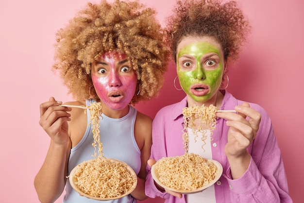 Verbaasde vrouwen met krullend haar passen schoonheidsmaskers toe op gezicht eten heerlijke pasta reageren op iets schokkends kijken horrorfilm terloops gekleed geïsoleerd over roze achtergrond Gezichtsbehandelingen