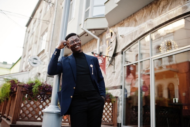 Verbazingwekkend uitziende Afro-Amerikaanse man draagt blauwe blazer met broche zwarte coltrui en bril op straat Modieuze zwarte man