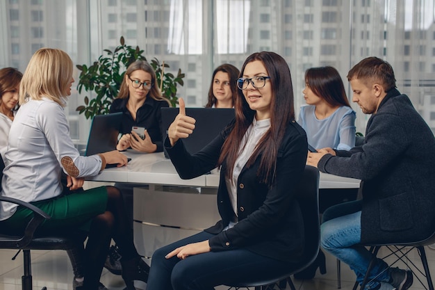 Verbazingwekkende charmante vrouwelijke werknemer met een bril die op een stoel op kantoor zit en de vinger opheft, omringd door haar collega's die ondergedompeld zijn in het werk. Concept van succes