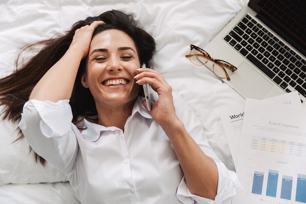 Verbazingwekkende mooie gelukkige jonge zakenvrouw in formele kleding binnenshuis thuis ligt op bed praten via de telefoon.