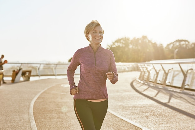 Verbetering van het gezondheidsportret van een gelukkig gemotiveerde volwassen vrouw in sportkleding die buiten op een zonnige dag loopt