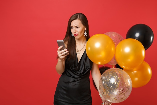 Verbijsterde jonge vrouw in kleine zwarte jurk met luchtballonnen met behulp van mobiele telefoon tijdens het vieren van geïsoleerd op rode achtergrond. Valentijnsdag Happy New Year verjaardag mockup vakantie partij concept.