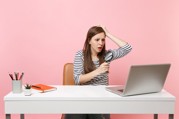 Verbijsterde vrouw die zich aan haar hoofd vastklampt en door vergrootglas op pc-laptop kijkt terwijl ze op kantoor aan een project werkt