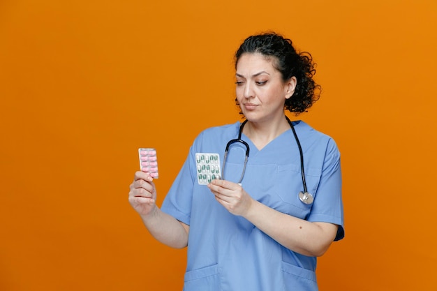 Verdachte vrouwelijke arts van middelbare leeftijd, gekleed in uniform en stethoscoop om de nek, met pakjes pillen die naar hen kijken geïsoleerd op een oranje achtergrond