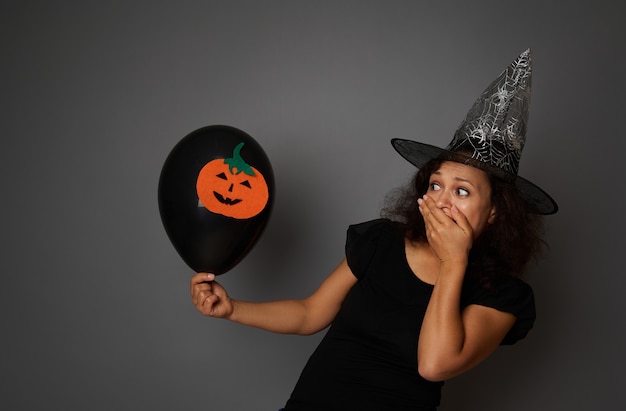 Verdoofde vrouw gekleed in zwarte heks carnaval kleding bedekt haar mond van afschuw, kijkt naar een zwarte luchtballon met vilt gesneden pompoen in de hand. halloween-concept met exemplaarruimte op grijze achtergrond