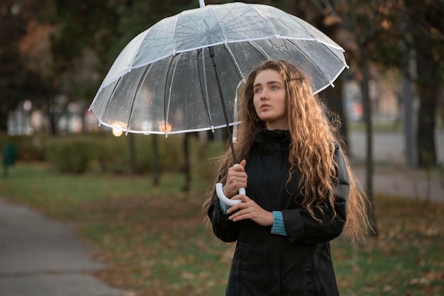 Verdriet aantrekkelijke vrouw met lang haar in herfstpark bij regenachtig weer onder transparante paraplu Portret van mooi meisje