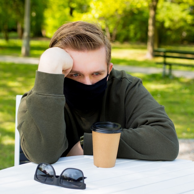 Verdriet Man in een beschermend zwart masker zittend aan een tafel in een café. Virusbescherming. Mannen in een masker koffie drinken in een stadspark. Kopieer ruimte.