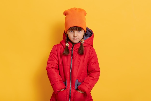 Verdrietig beledigd meisje dat een oranje pet en een rood jasje draagt dat zich tegen de gele muur bevindt en naar de voorkant kijkt met een verstoorde gezichtsuitdrukking