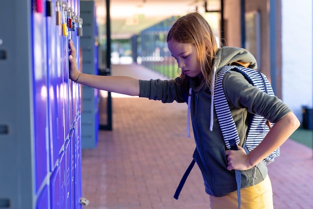 Verdrietig blank schoolmeisje met schooltas leunend op kastjes op school met kopieerruimte