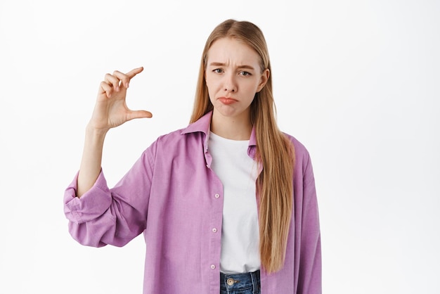 Verdrietig en teleurgesteld blond meisje dat een klein klein ding met de hand laat zien en fronsend van streek ontstemd door iets dat tegen een witte achtergrond staat