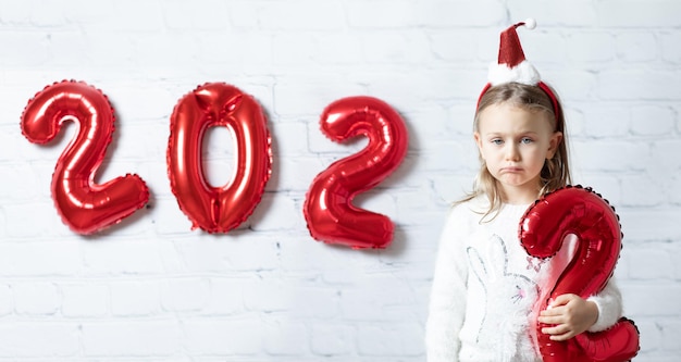 Verdrietig kind in kerstmuts geïsoleerd wit pauze muur achtergrond cijfers ballonnen nieuwjaar nummers 2022