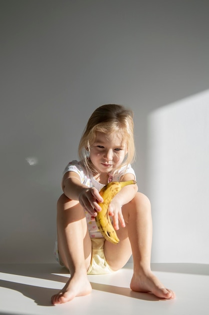 Verdrietig kind meisje blond met een banaan zittend op de vloer tegen de achtergrond van een witte muur