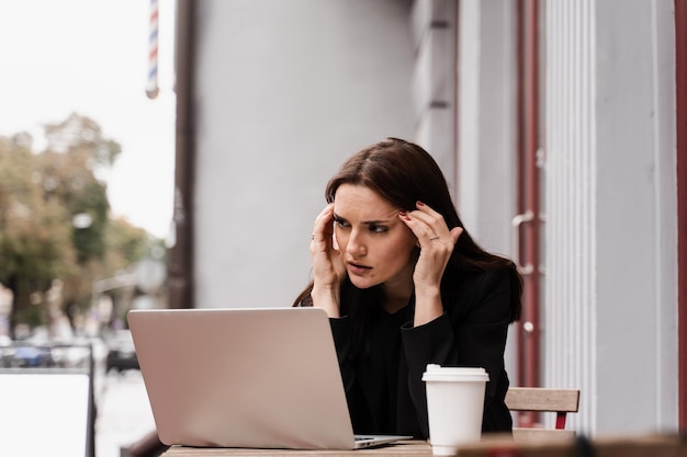 Verdrietig meisje met laptop en kopje koffie kreeg migraine raakt haar hoofd vanwege pijn Vrouw manager met sterke hoofdpijn die op afstand online op laptop werkt buiten café