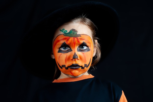 Verdrietig meisje met pompoenmake-up op haar gezicht voor halloween, in een zwarte hoed, eenzaamheid en verdriet op vakantie.