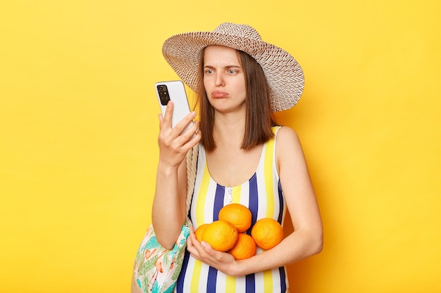Verdrietig overstuur ontevreden vrouw met gestreept zwempak en hoed geïsoleerd op gele achtergrond fruit vasthoudend met behulp van mobiele telefoon kijkend naar smartphonescherm met ongelukkige uitdrukking
