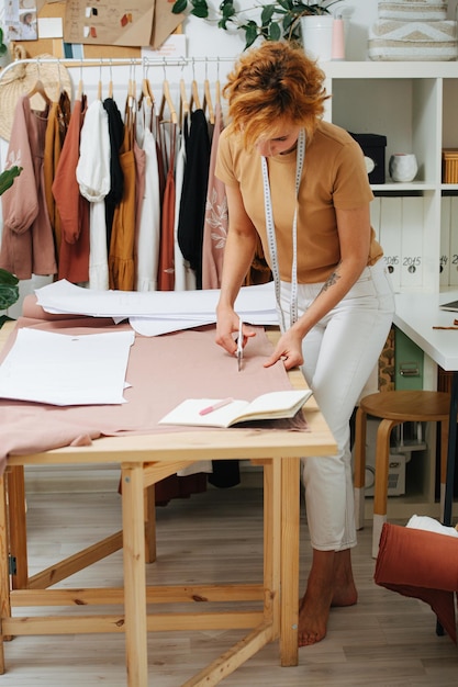 Verdwaald in haar werk jonge naaister die een stof op tafel knipt Meter hangt aan haar nek Planken en rekken achter Ze heeft kort gember geverfd haar en kijkt naar beneden