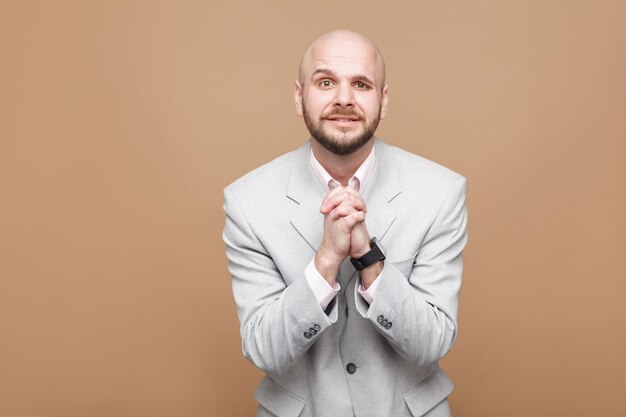 Vergeef me alsjeblieft boos. Kale, bebaarde zakenman van middelbare leeftijd in een klassiek lichtgrijs pak dat staat en naar de camera kijkt met een hoopvol gezicht. indoor studio opname, geïsoleerd op bruine achtergrond.