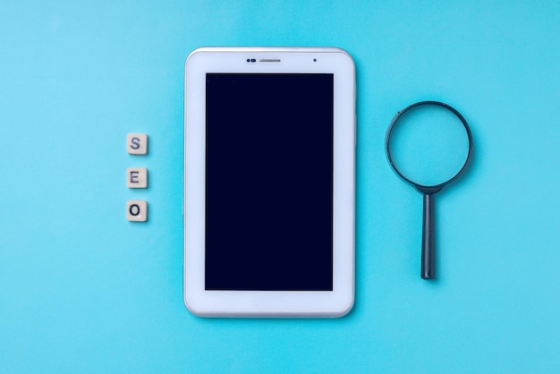Vergrootglas, tablet-smartphone en houten kubus belettering SEO op blauwe achtergrond.