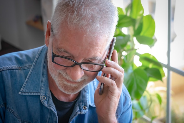 Vergrote weergave van een senior volwassen man met wit haar en baard met behulp van de mobiele telefoon - Intens licht uit het raam. Een persoon met een spijkerblouse.