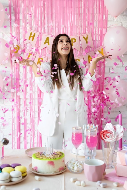 Verjaardagsfeestje Verjaardagstafels Aantrekkelijke brunette vrouw in witte feestkleding die verjaardagstafel voorbereidt met taarten, cakepops, macarons en andere zoetigheden die roze confetti gooien