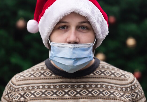 Verkeerd dragen van een masker. Close-up portret van man met een kerstman hoed, xmas trui en medische masker met emotie. Tegen de achtergrond van een kerstboom. Coronapandemie