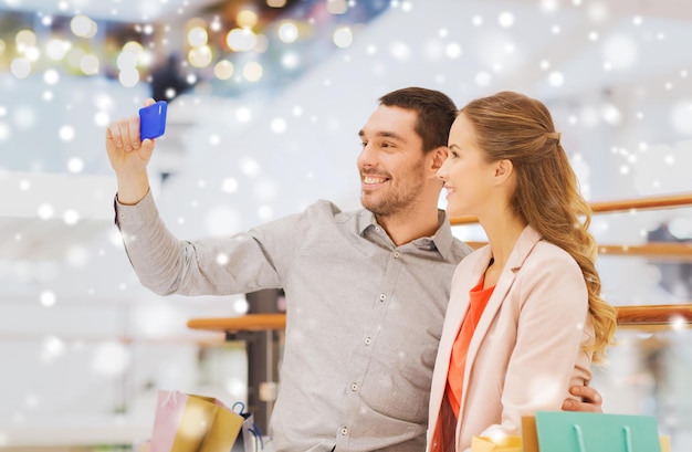 verkoop, consumentisme, technologie en mensenconcept - gelukkig jong stel met boodschappentassen en smartphone die selfie nemen in winkelcentrum met sneeuweffect