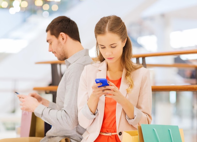 verkoop, consumentisme, technologie en mensenconcept - jong stel met boodschappentassen en smartphones in winkelcentrum